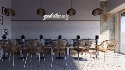 Ентериер на кафуле со бели ѕидови од тула, ратан столици и неонски натпис „good vibes only“.